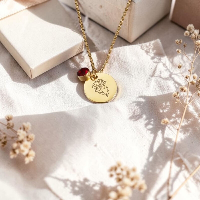 Gold necklace with a floral pendant and red birthstone on a light surface with dried flowers.